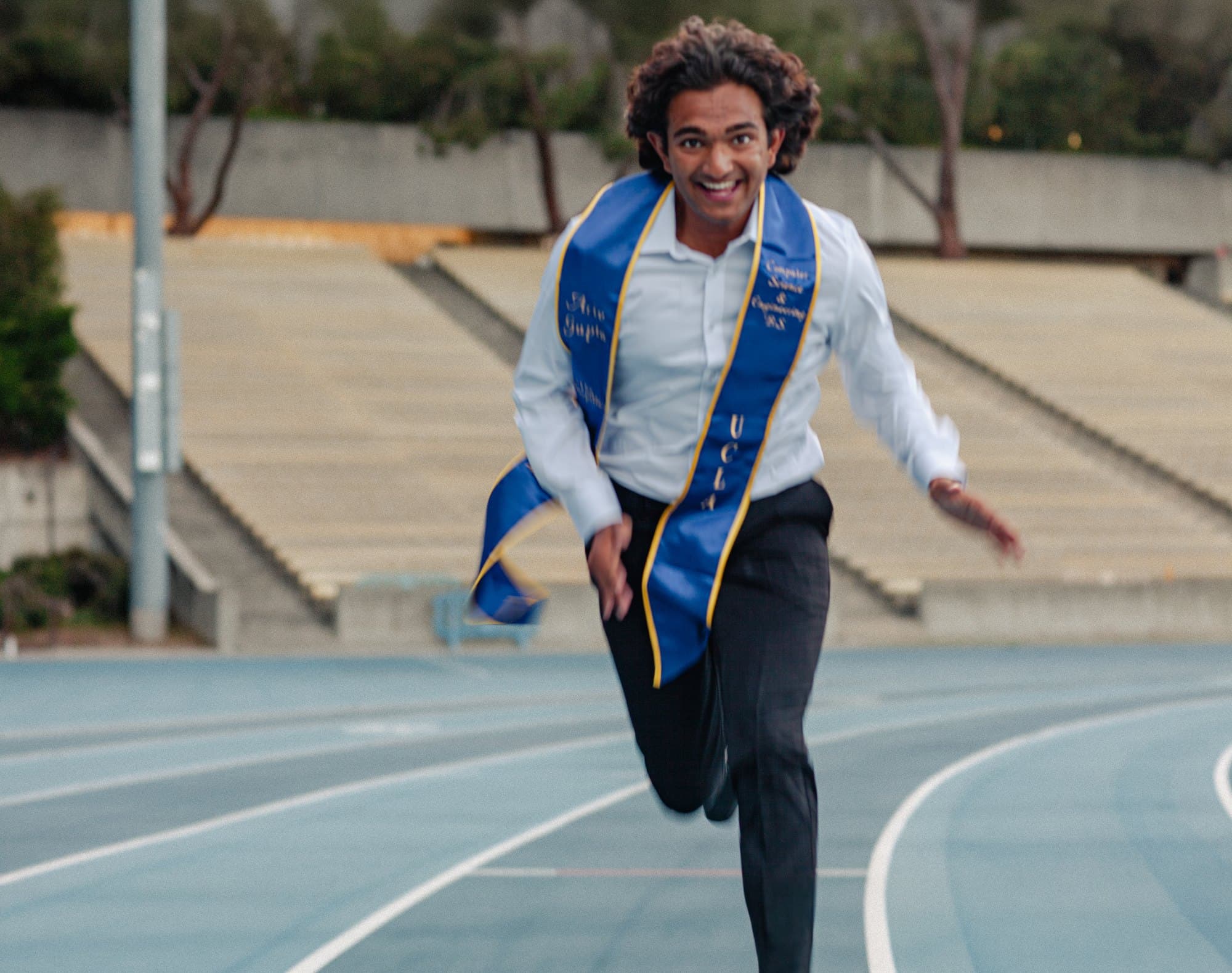 Ariv sprinting down a track in his UCLA graduation stole, mid-laugh