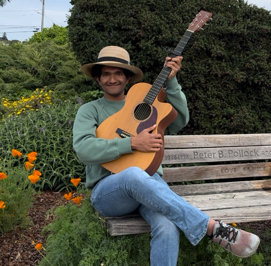 Ariv on a garden bench in a straw hat, holding his acoustic guitar among the poppies